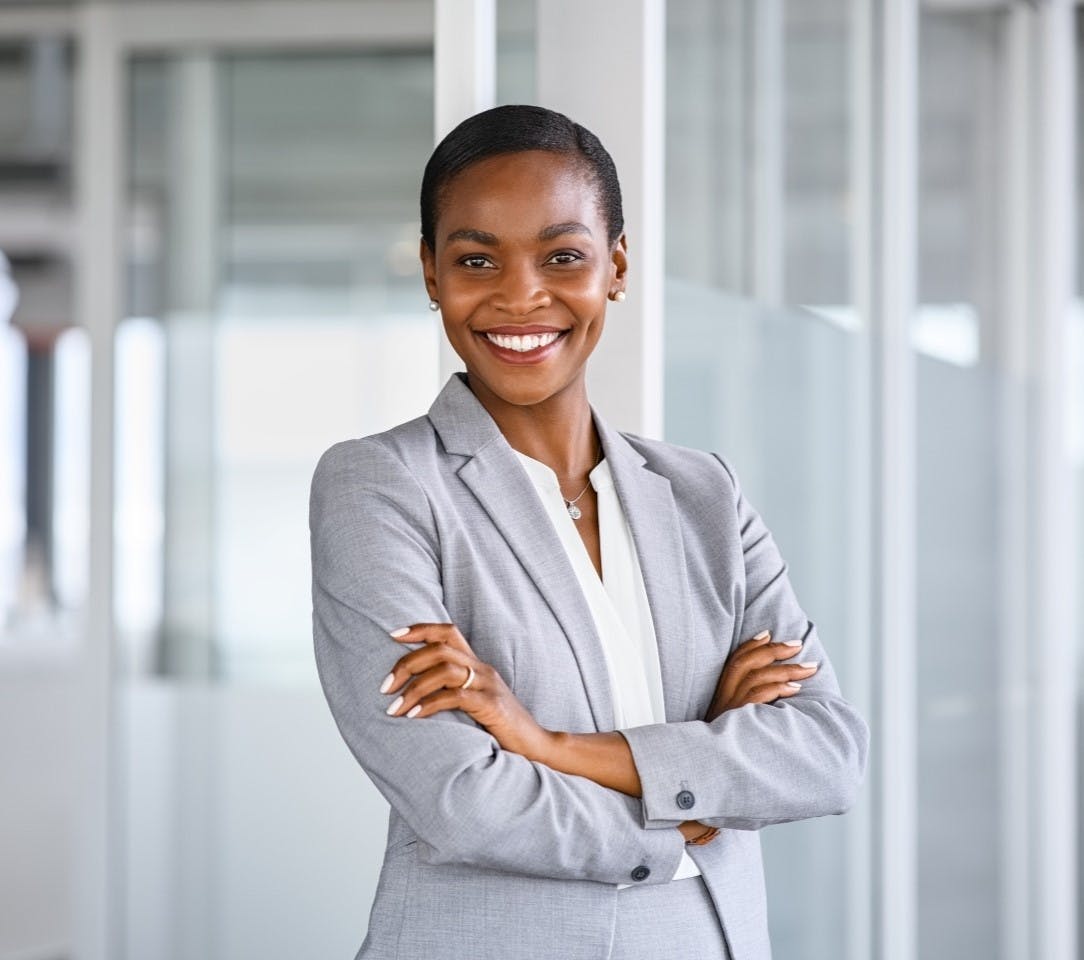 A woman in a grey blazer smiles confidently with her arms crossed, standing in a modern office setting.