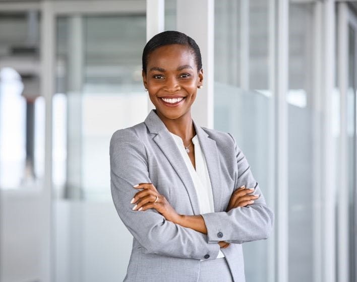 A woman in a grey blazer smiles confidently with her arms crossed, standing in a modern office setting.