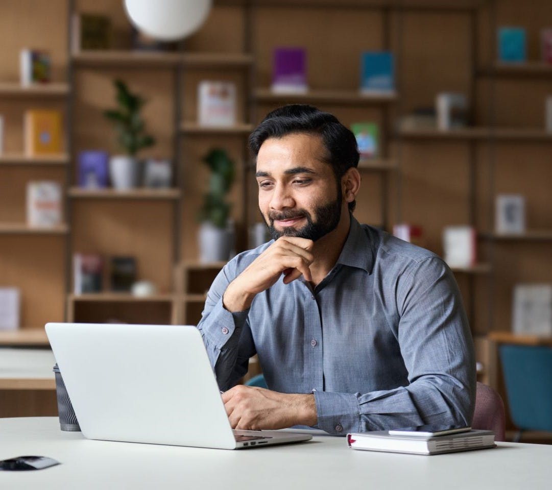 Smiling Indian businessman working on laptop in modern office lobby space.