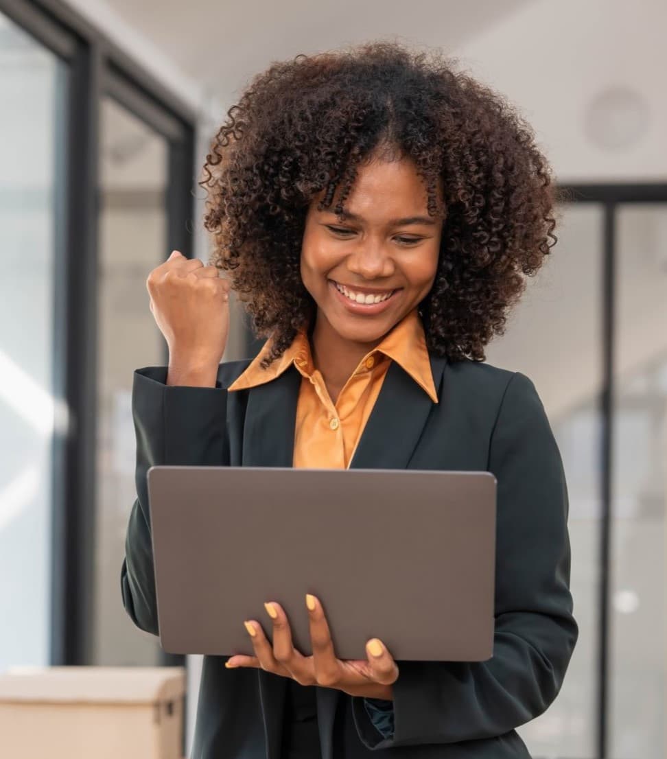 Beautiful African American female wearing an orange shirt , looking at her laptop celebrating and smiling.