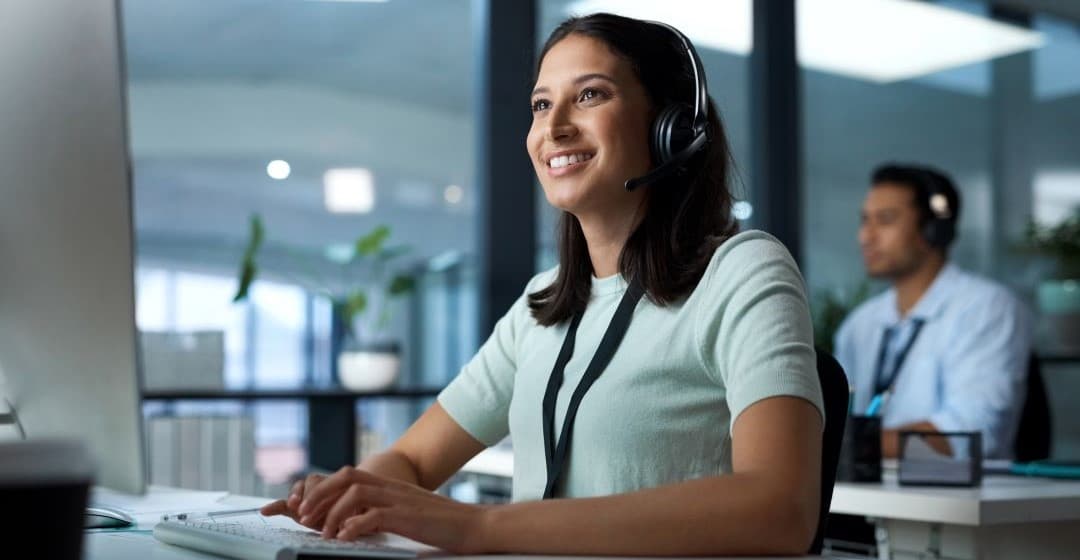 Smiling dark haired female typing on computer wearing a headset.