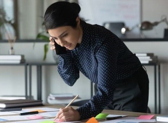 Serious Indian businesswoman talking on her business phone number, writing notes on colorful sticky papers in office.