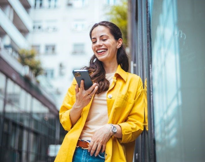 Brunette female wearing a yellow blazer smiling at her phone.