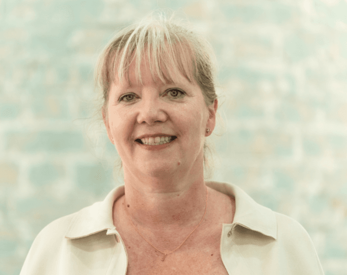 Portrait of smiling blonde female telephone answering Chief Operating Manager Ceri Henfrey on a blue brick background.