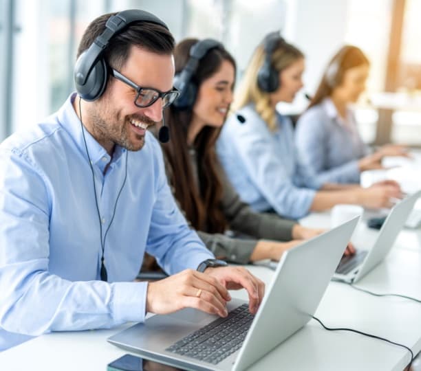 Happy male live chat operator with headset and laptop, chatting with client. Group of colleagues working on laptops in the background.