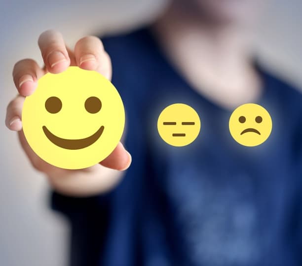 A person holding an illustrated smiley face with a neutral and sad expression illustrated beside it, showing customer satisfaction