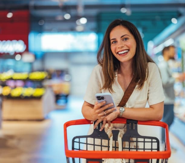 Image of a happy customer leaning on a shopping trolley in a supermarket with a phone in her hand, smiling, representing customer lifetime value